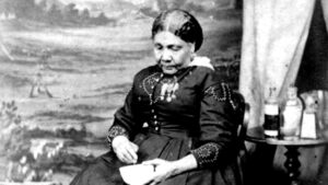 Photograph of Mary Seacole seated in front of a wide valley, beside a tent, next to a camp stool and table full of medicine bottles. Mary is wearing a military looking outfit thought to have been self-designed. This is the last picture of Mary, taken around 1873. The reason for its commission is unknown